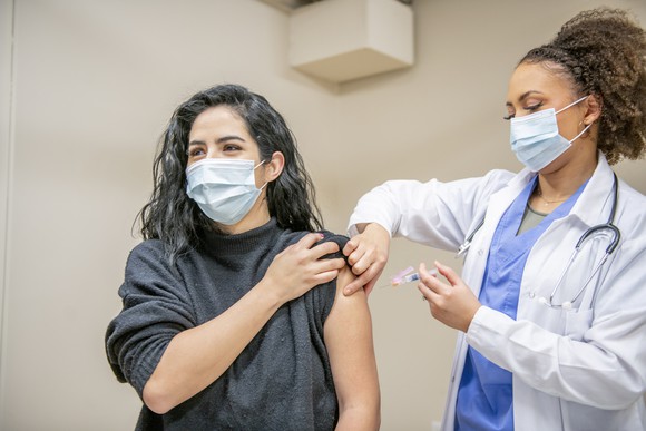 A masked healthcare professional holding a syringe with needle near a masked patient's arm.