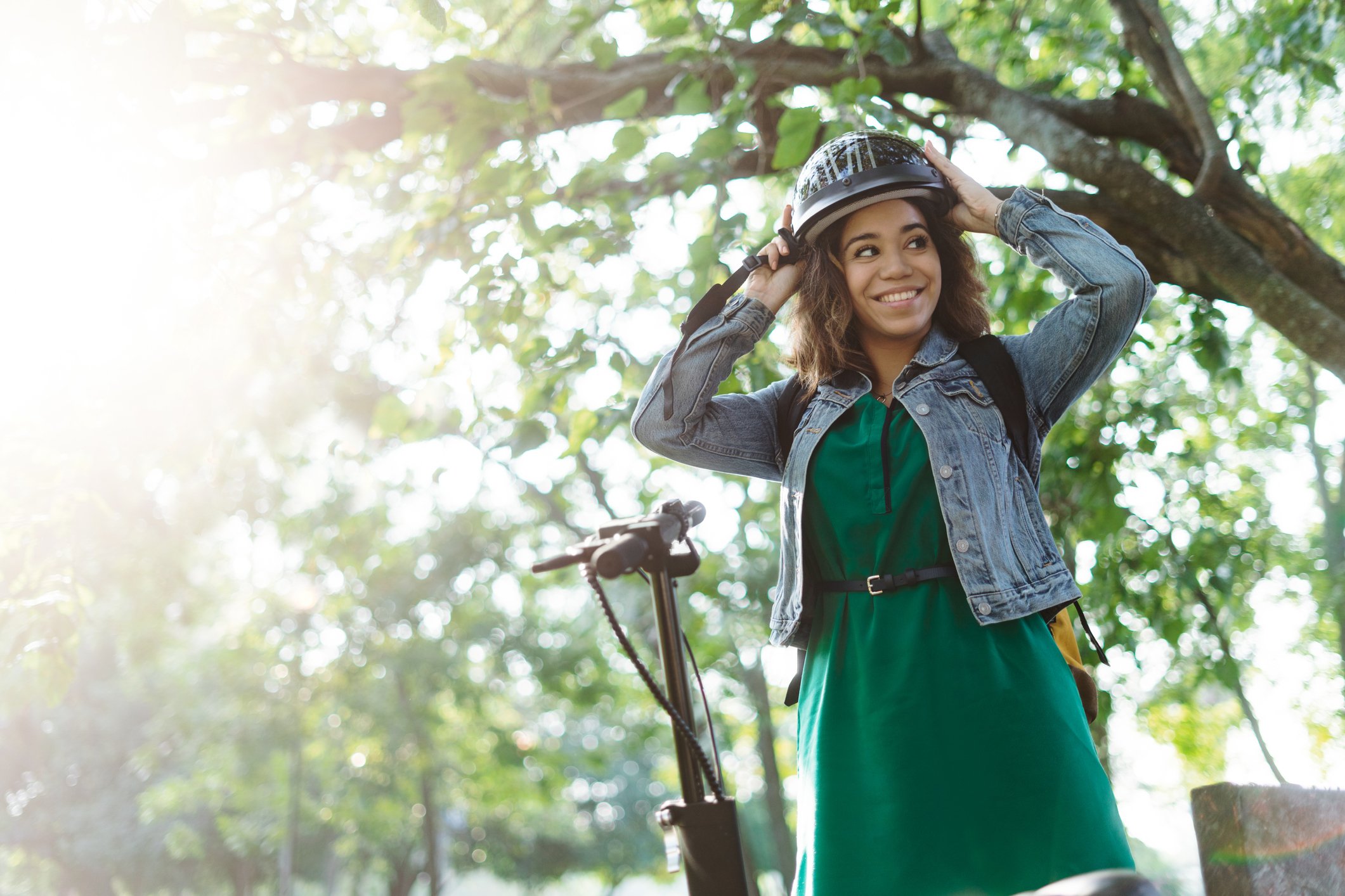 A smiling woman stands next to her scooter and adjusts her helmet.