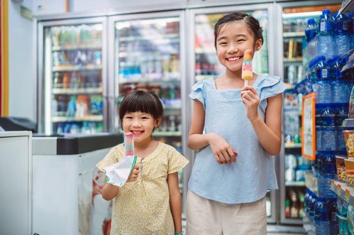 Two young children smile and enjoy popsicles in front of convenience store refrigerators.