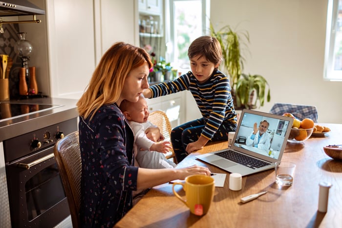 A mother holding a baby sits next to her other child at the dinner table while the three consult with a telemedicine doctor via a laptop.