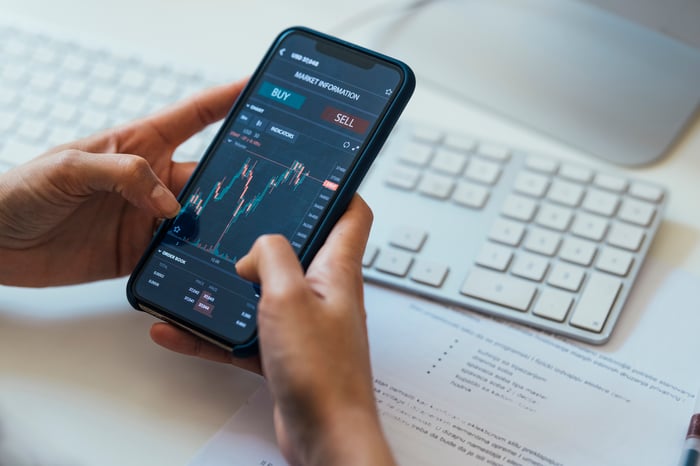 A person holding a smartphone that's displaying a stock chart, along with buy and sell buttons.