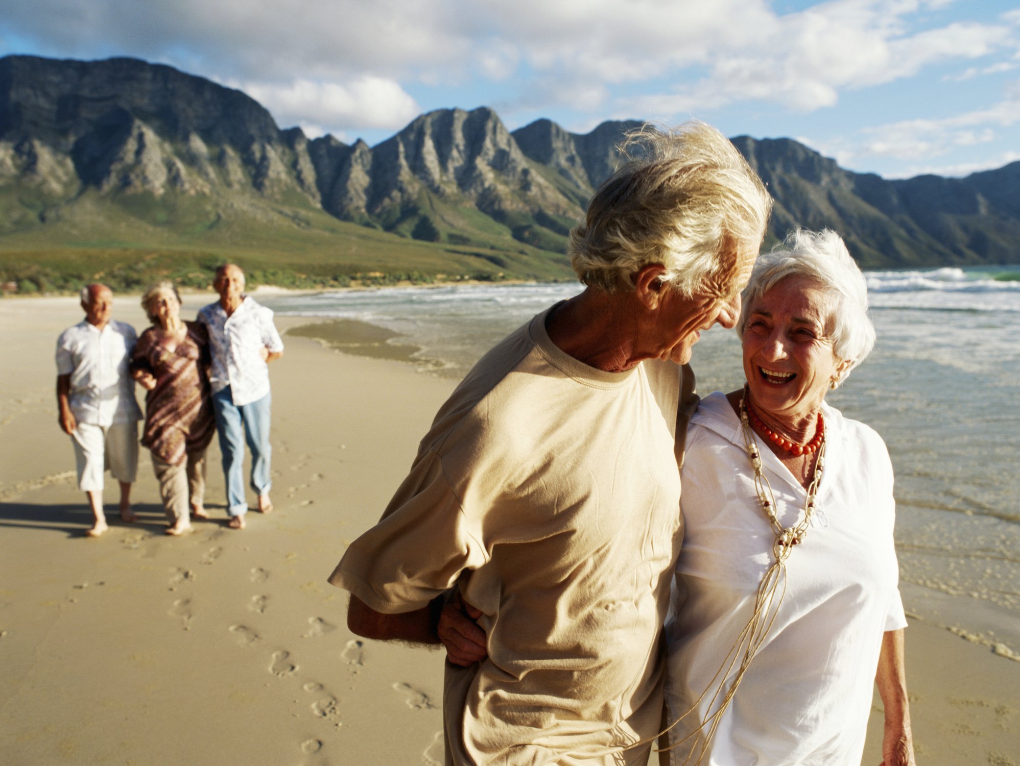 couple walking in foreground along beach smiling with mountains in background