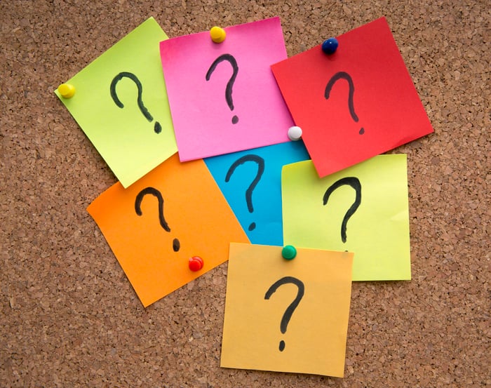 Seven square pieces of paper of various colors, with a question mark written on each, tacked to a bulletin board.