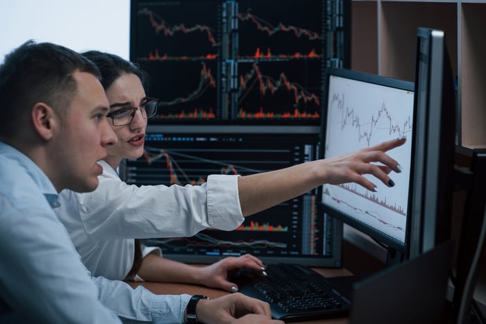 Two persons appearing confused while studying stock price charts on computer screens.