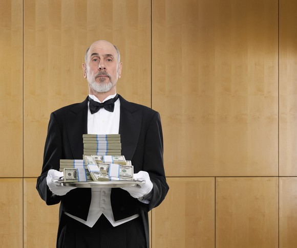 A butler in a tuxedo holding a tray full of cash.