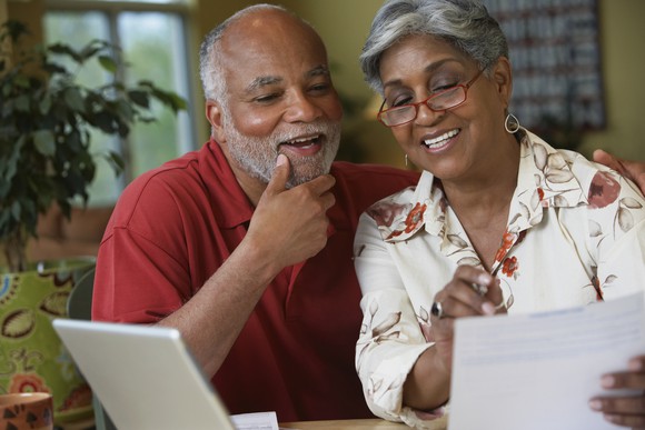 Senior couple smiling while reviewing paperwork at home in front of laptop.