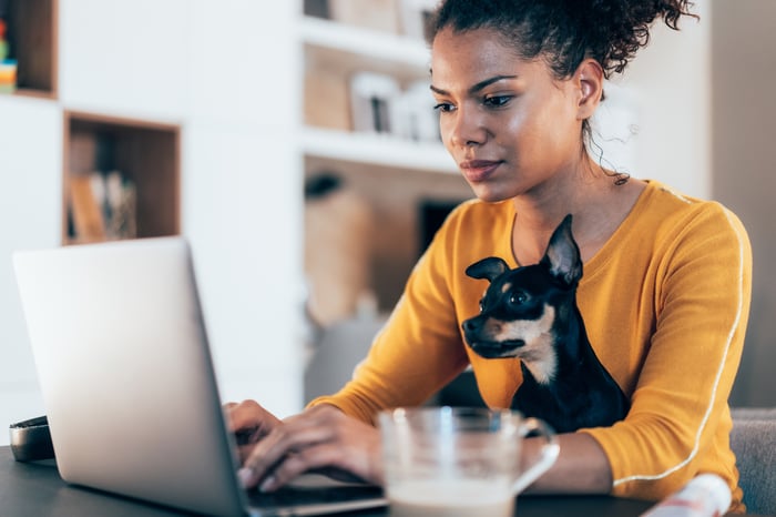 A woman typing on a laptop, with a small dog seated on her lap. 