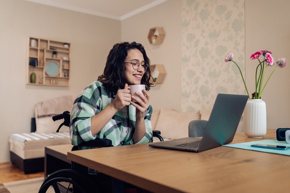 A woman in a wheelchair holding a coffee mug while looking at an open laptop.
