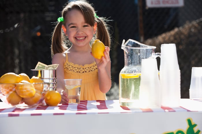 A girl holding a lemon and standing in front of a table with lemonade, cups, and lemons.