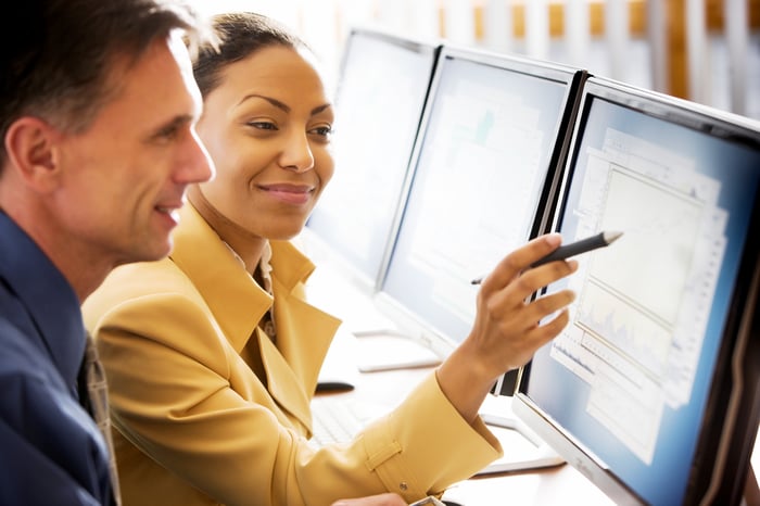 Two people smiling while looking at multiple computer monitors.