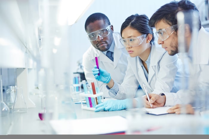 Three lab technicians examining liquid in test tubes and making notes.