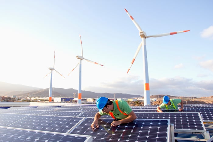 Workers inspecting solar panels with wind turbines in the background.