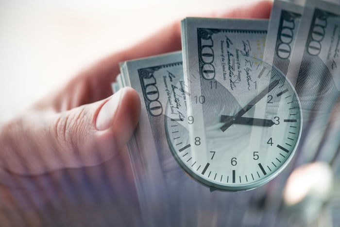 A clock superimposed on a fanned stack of one hundred dollar bills.  