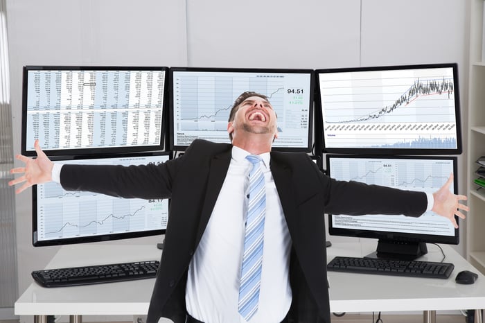 A stock broker celebrating with arms open wide and screens behind him displaying rising stock price charts.