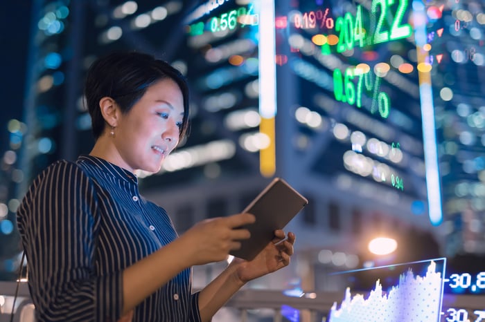 An businessperson checking a tablet in front of a stock market display board.