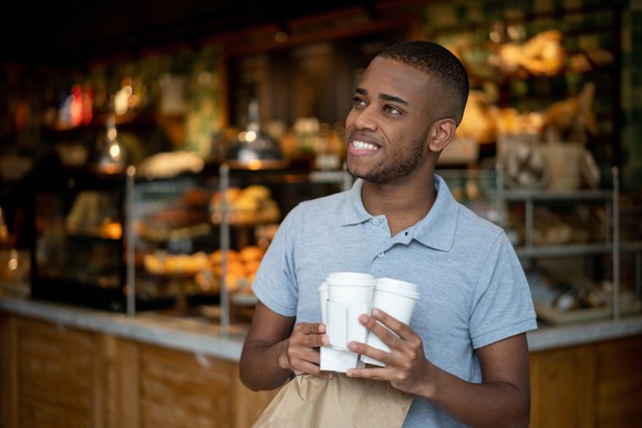 A person holding cups of coffee and a takeout bag walks away from a coffee shop counter.