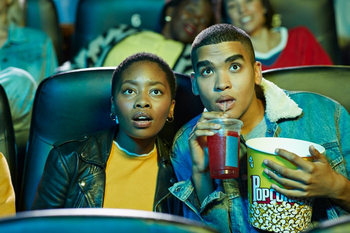 A young couple eating popcorn and drinking a beverage while watching a movie in a theater.