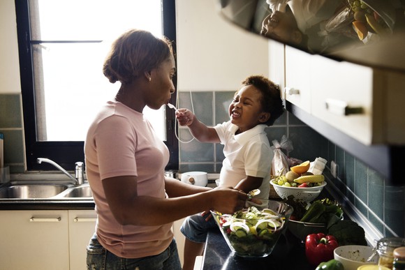 An adult prepares a salad in the kitchen with a young child smiling and sitting on the counter.