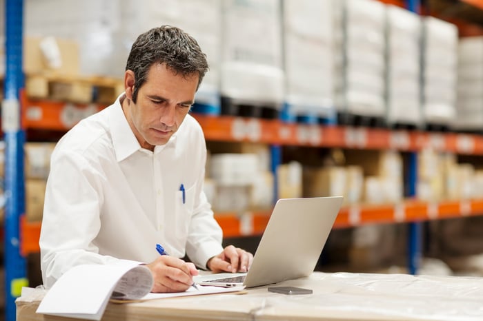 A person in a warehouse using a laptop and writing on a clipboard.