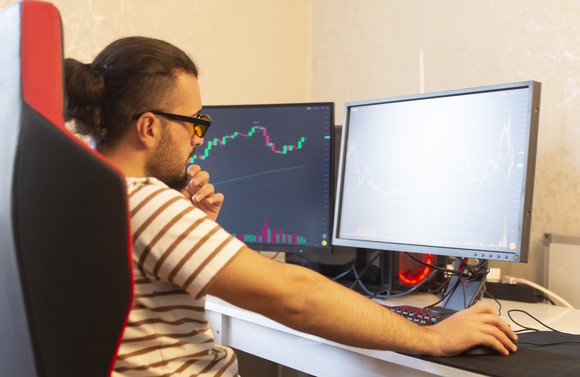 A person sits at their computer monitoring two screens, one of which shows a stock chart.