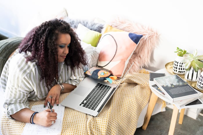 Person lying on bed writing note and looking at laptop