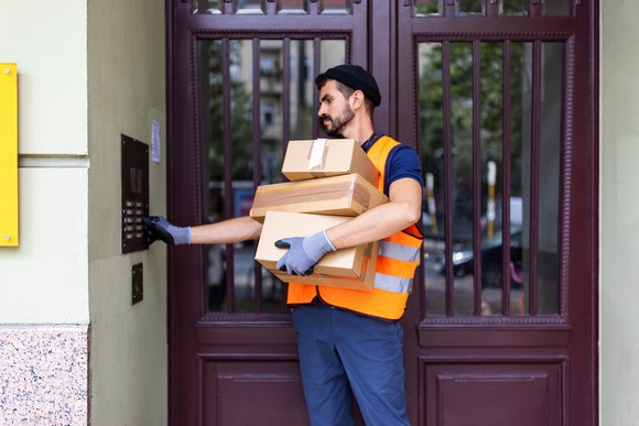A delivery person ringing a doorbell to make a delivery while holding three boxes.