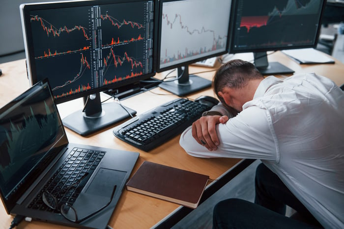 A businessperson resting his head on table with computer screens in front displaying crashing stock price charts.