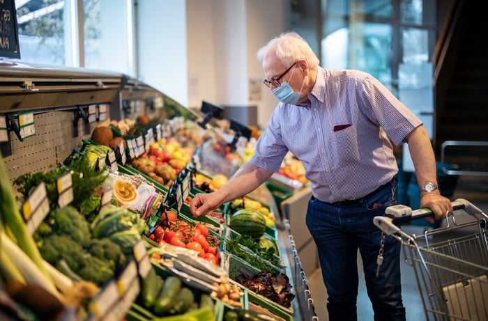 A senior person wearing a mask and picking vegetables at a grocery store.