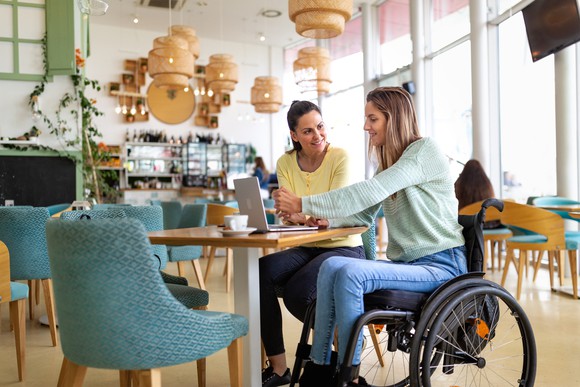 Two people, one in a wheelchair, using a laptop in a cafe.