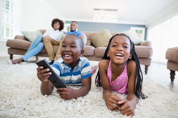 Two siblings lying on a rug watching television, with their parents seated on a couch in the background.