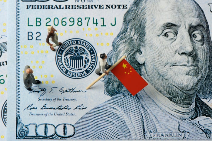 On a $100 bill Benjamin Franklin looks worriedly over his shoulder at people carrying a Chinese flag