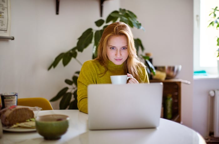 Person sitting at a desk holding a mug and looking at a laptop.