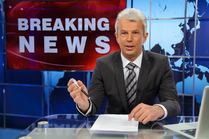 A news anchor in a studio with breaking news flashing behind on screen.