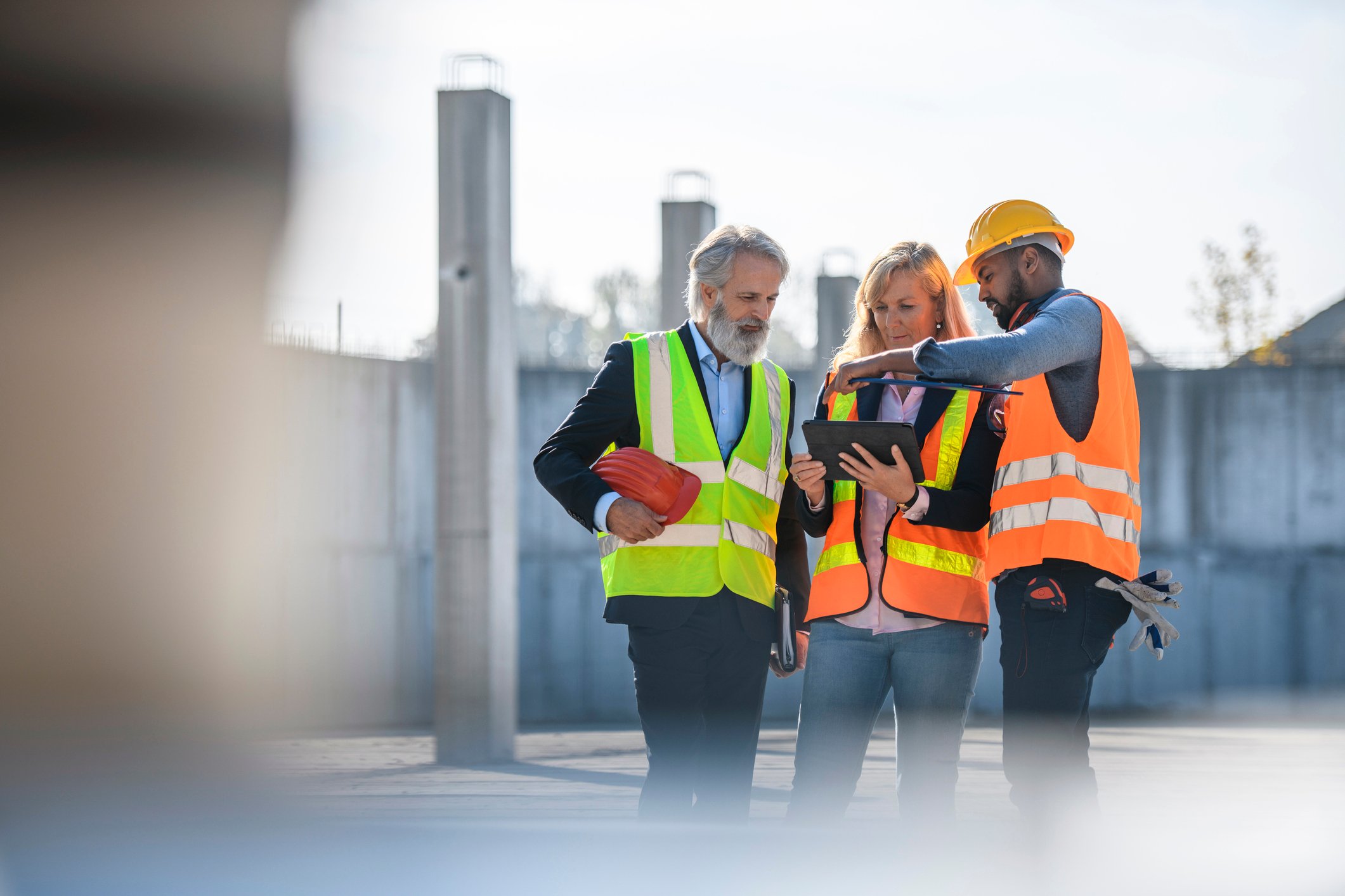 Three people wearing bright working vests discuss a matter while looking at a tablet onsite.