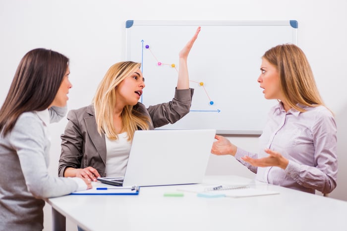 Three business professionals arguing in a conference room with a stock chart in the background.