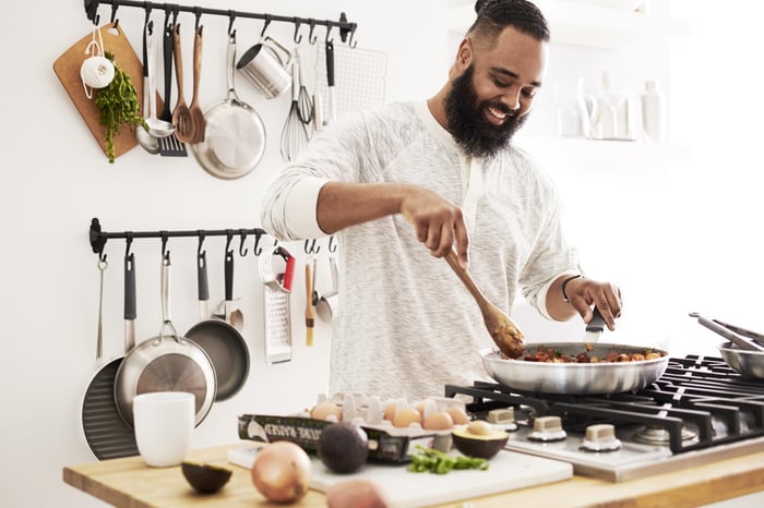 A man cooking on a stovetop, with various kitchen implements hanging in the background.