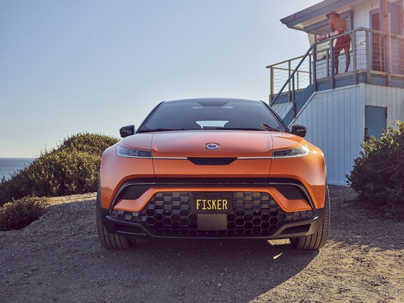 Front view of an orange Fisker Ocean SUV parked near a seaside house.