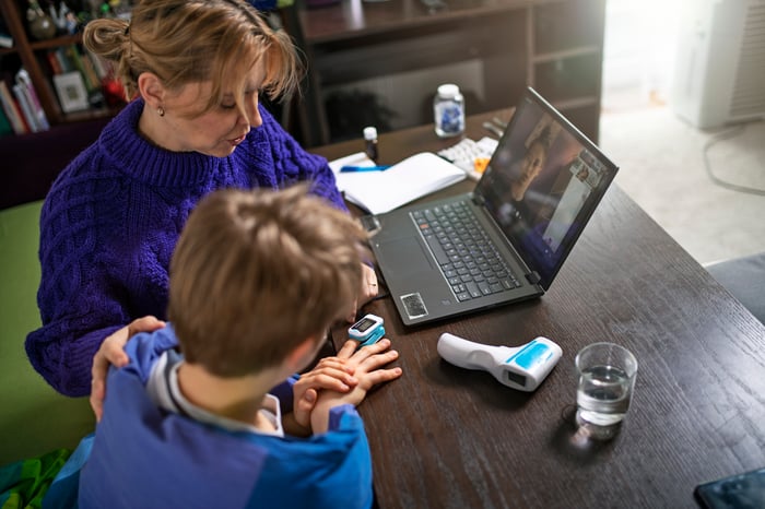 An adult and child attend a telehealth appointment via a laptop, and the child is wearing a pulse oximeter.