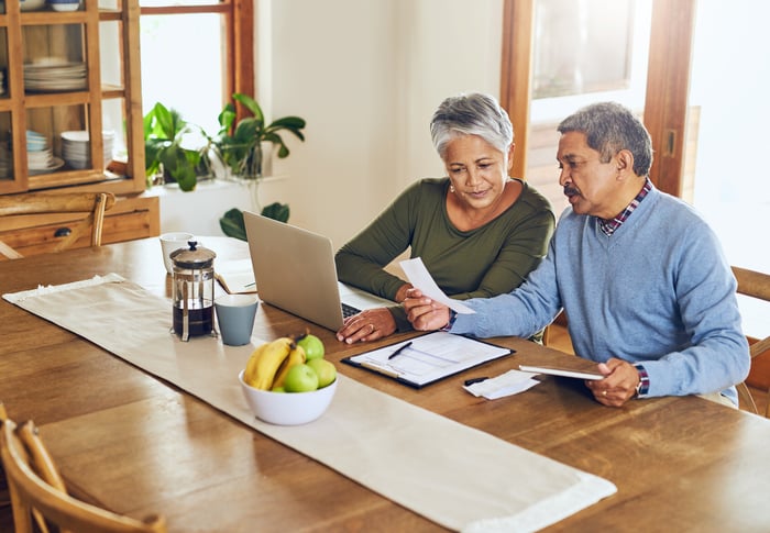 Two older adults sitting at a table looking at a laptop screen and paperwork.