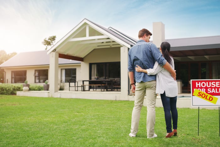 A couple standing in front of their new home, with a sold sign out front