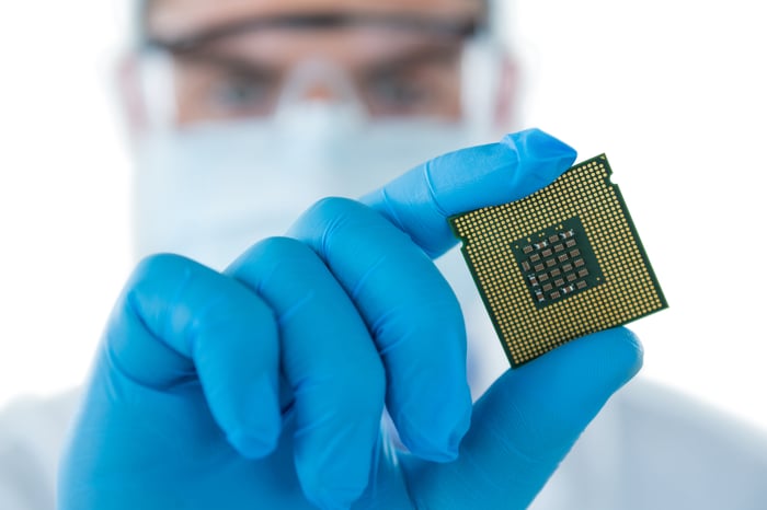 A person in a lab wearing protective clothing, holding a computer chip in their gloved hand