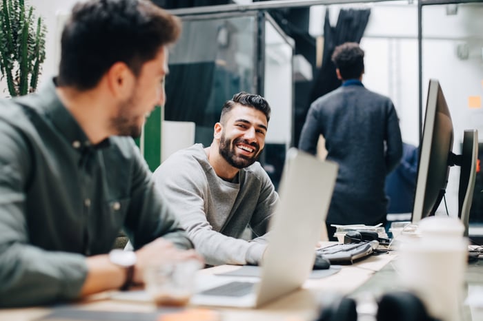 One seated colleague smiling at another at their computers in their office workspace.