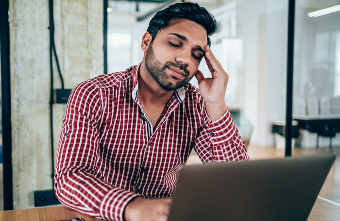 A businessman holds his head while working on a laptop.