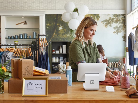 A point-of-sales device from Square sits on a counter in a retail location. A person is standing next to it.