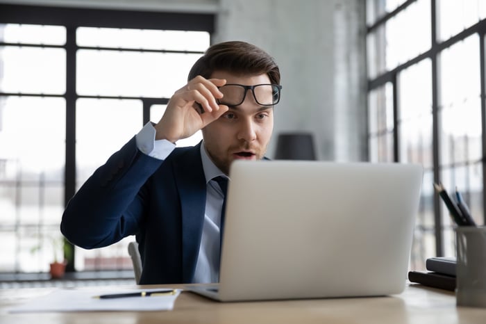 A man lifts their glasses in surprise while reading information on a computer.