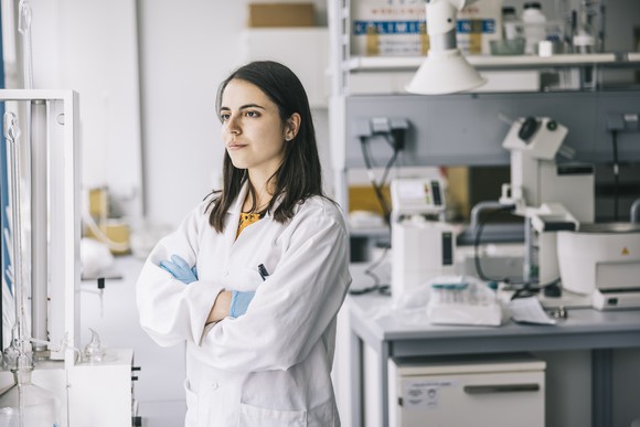 A scientist stands in her laboratory, looking away from her bench while thinking.