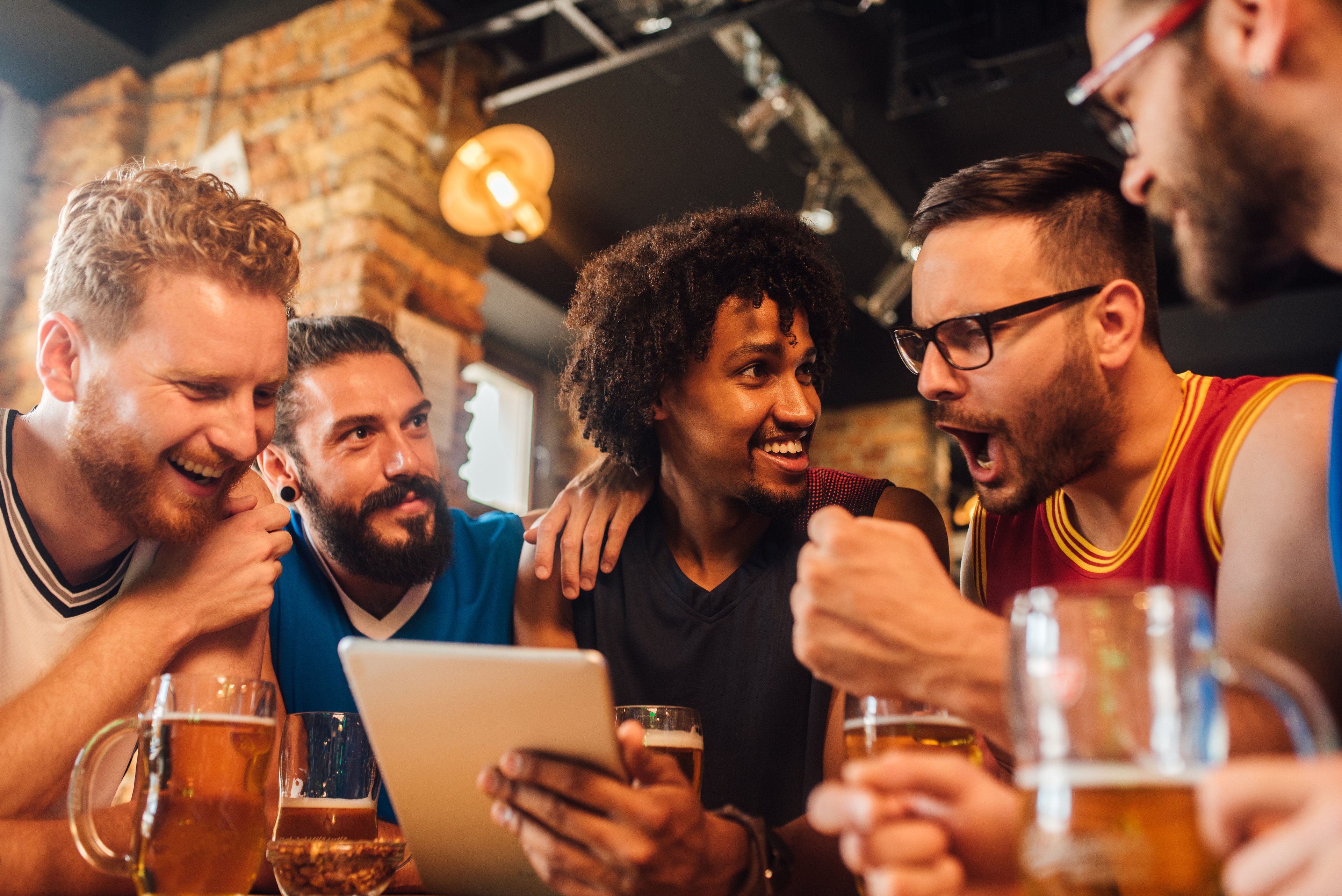 A group of cheering friends at a bar looking at a tablet device, placing online sports bets