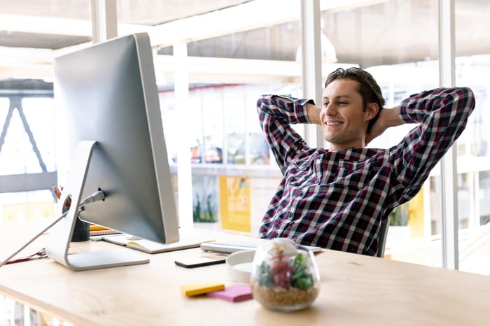 Man with hands behind his head smiling and looking at his computer monitor.