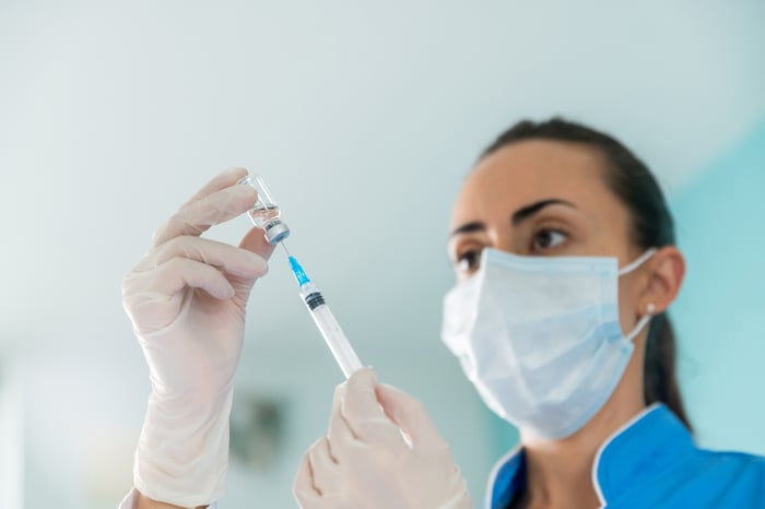 A masked healthcare worker draws up a dose in a hypodermic needle.