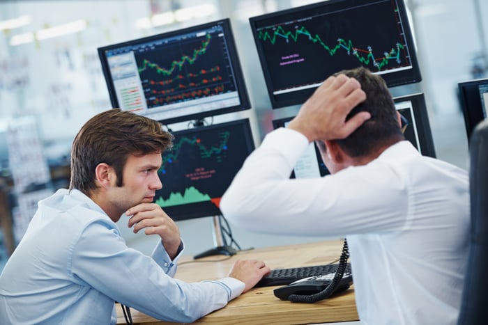 Two concerned persons looking at monitors displaying volatile stock price charts.